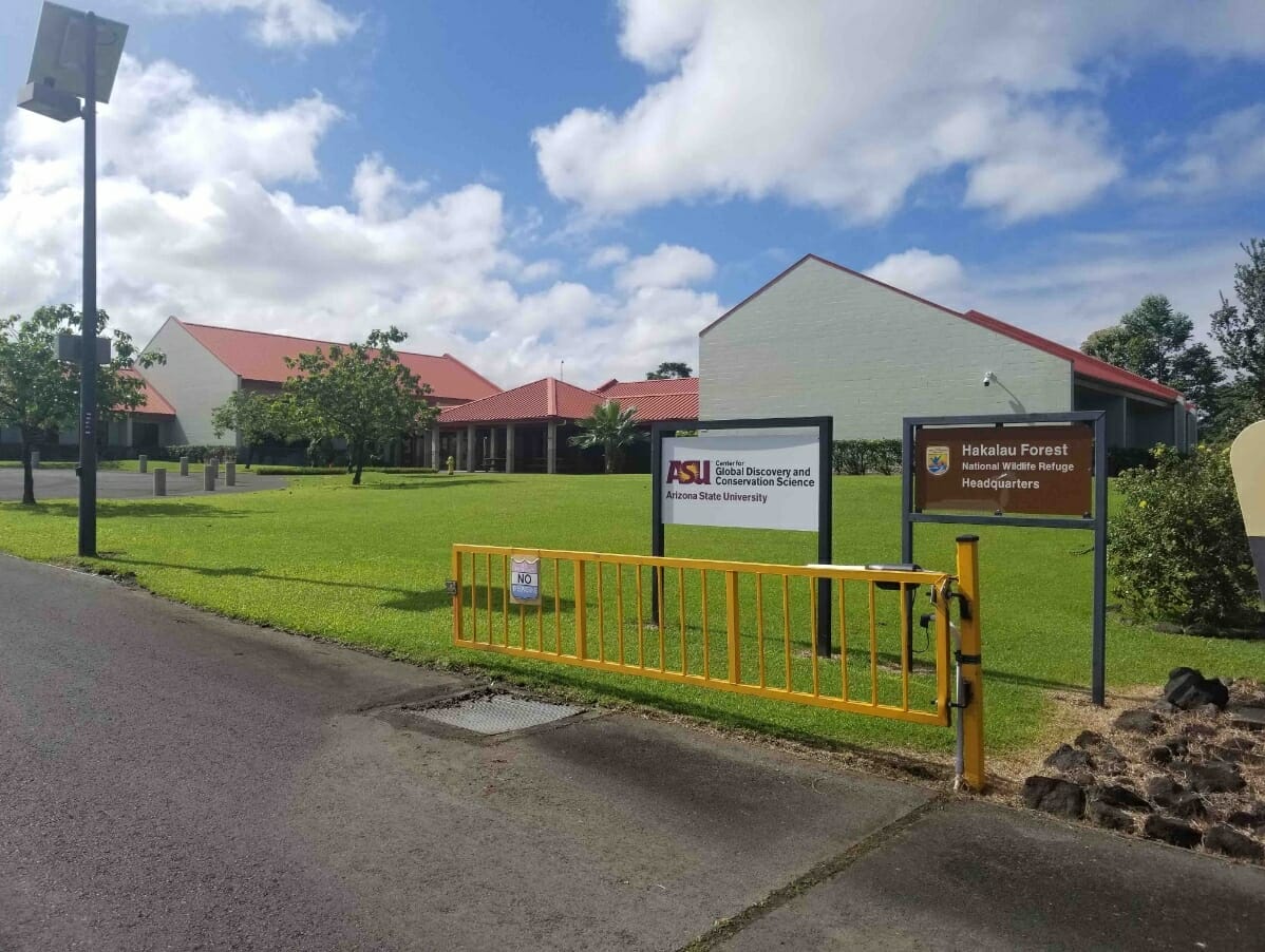 An image of the GDCS-Hawai’i headquarters, a welcoming sage green building with a red roof and verdant lawn, on a partly cloudy day.