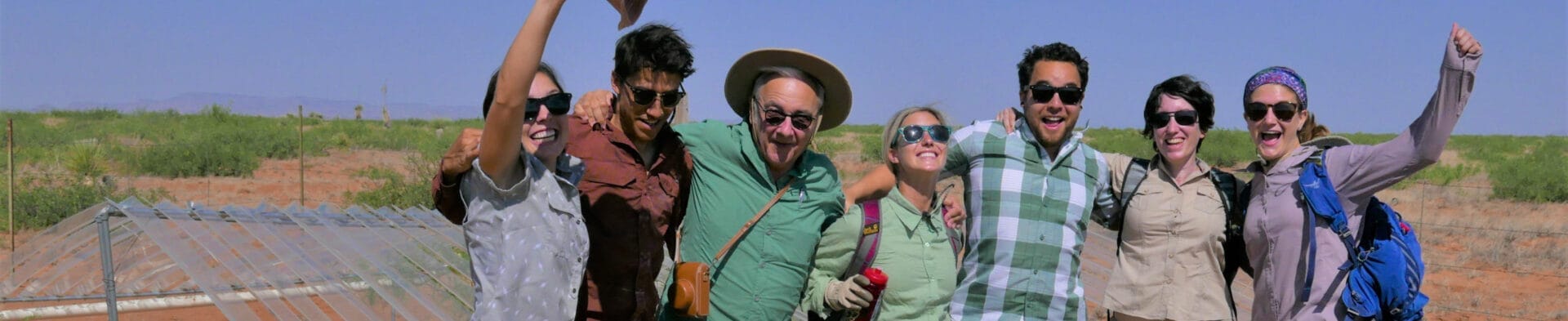 
		A group of seven people wearing sunglasses, smiling and laughing with their arms around each other. A desert landscape is visible in the background.		