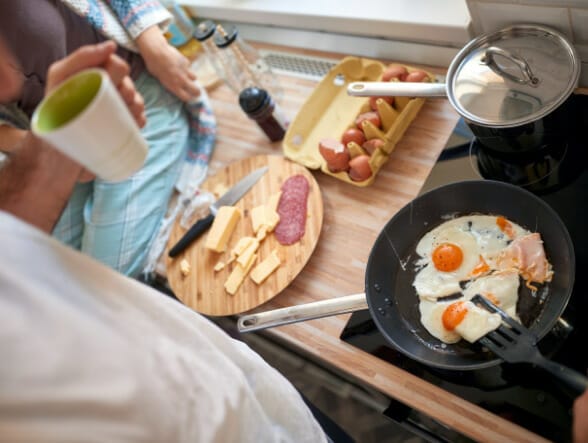 A kitchen scene - eggs frying in a black pan with an open egg carton and charcuterie board with sliced meat and cheese to the left and a person sitting on the counter mostly out of frame