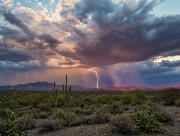 Ominous storm clouds hover over an early evening desert landscape with patches of sunlight in the foreground and lightning striking a mountain range in the distance